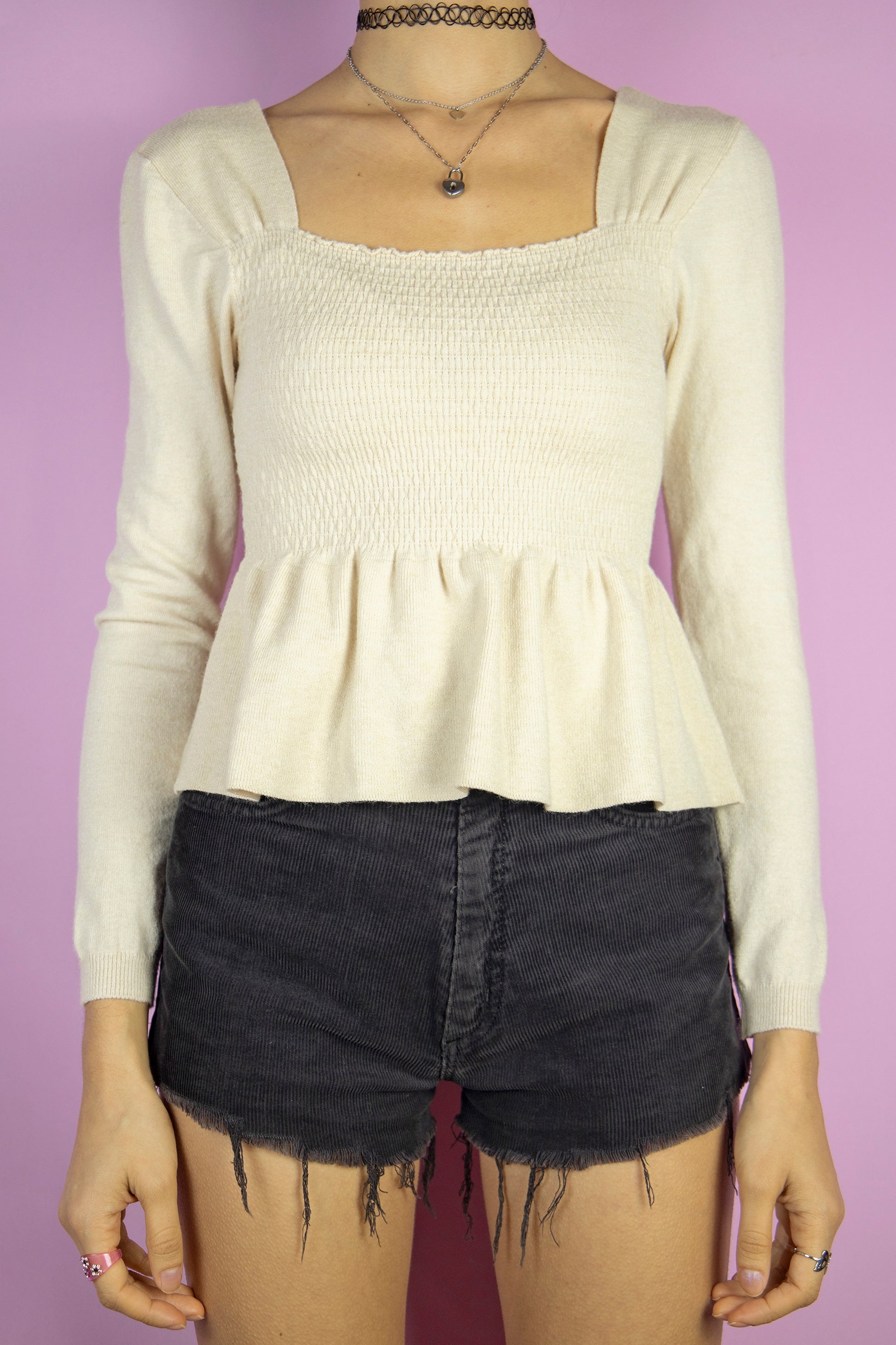 The Y2K Beige Shirred Peplum Top is a vintage 2000s knit blouse featuring a square neckline, long sleeves, and a shirred bodice that flows into a peplum hem.
