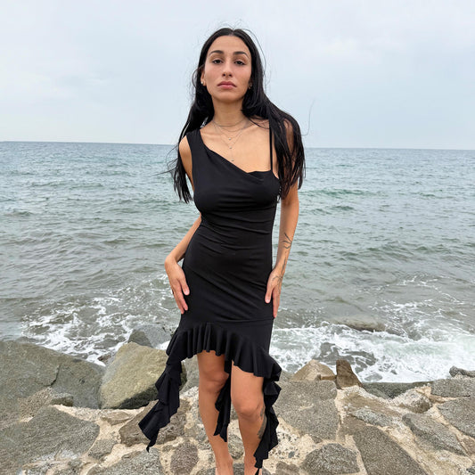 This vintage 2000s black mini dress features asymmetric straps with a subtle cowl neckline and a dramatic ruffled hem. Crafted from a stretchy fabric, it flatters the body with a bold party silhouette. Perfect for summer nights, club looks, or diva-inspired Y2K styling, this piece captures iconic 2000s glamour. Excellent vintage condition.