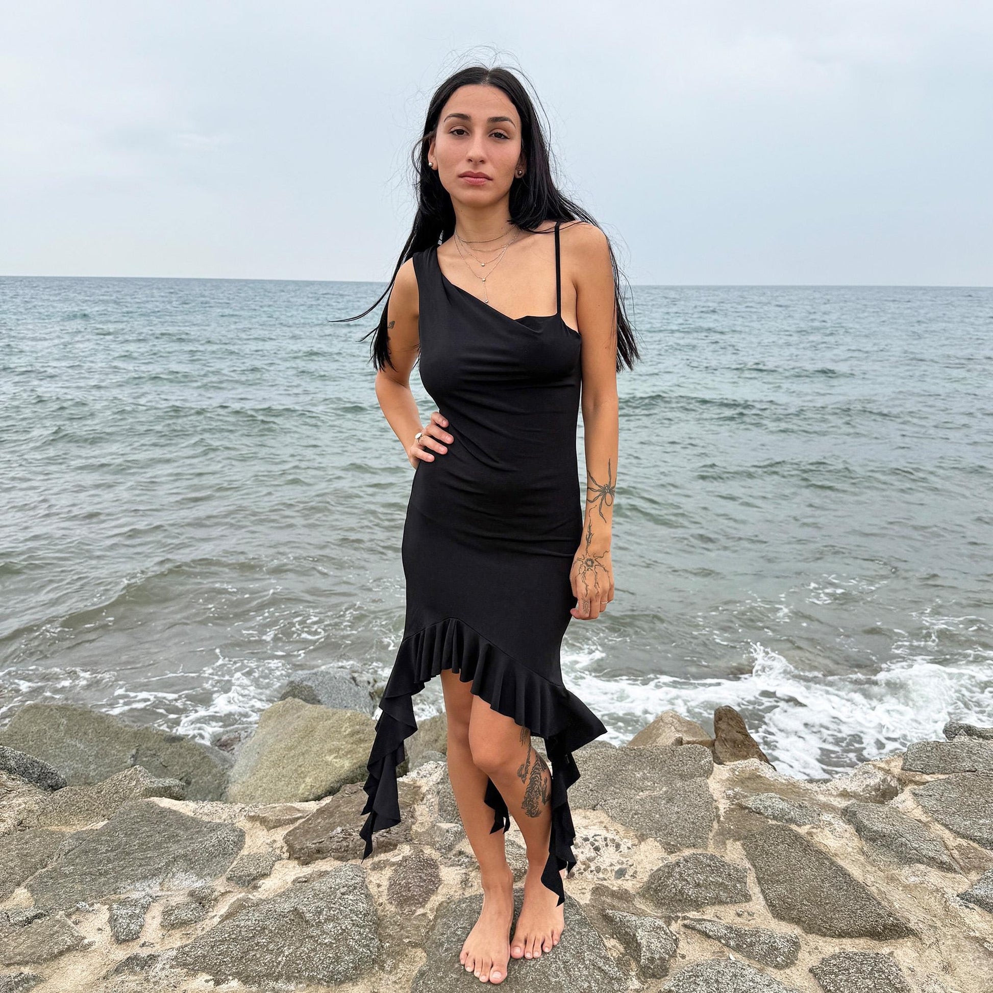 This vintage 2000s black mini dress features asymmetric straps with a subtle cowl neckline and a dramatic ruffled hem. Crafted from a stretchy fabric, it flatters the body with a bold party silhouette. Perfect for summer nights, club looks, or diva-inspired Y2K styling, this piece captures iconic 2000s glamour. Excellent vintage condition.