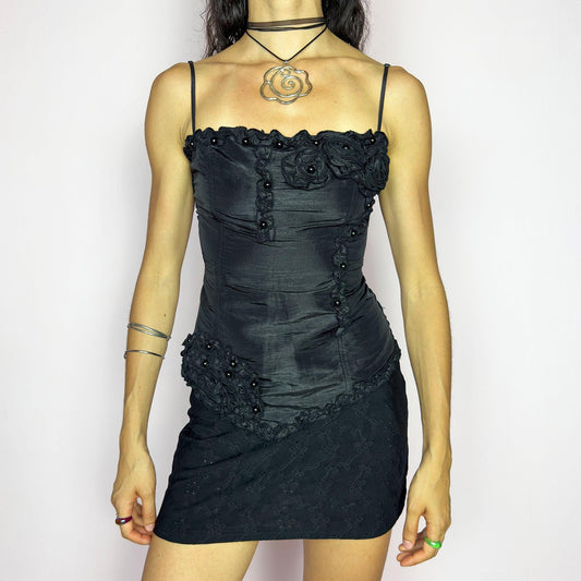 This vintage 2000s black corset top features adjustable spaghetti straps, floral rosette appliqué, beaded trim and an asymmetrical hem. The structured front sculpts the waist, while the ruched smocked back adds stretch and comfort. Ideal for prom, weddings, or dreamy nights out. A romantic Y2K formal top that blends fairytale opulence with a bold, sculptural silhouette. Great vintage condition.