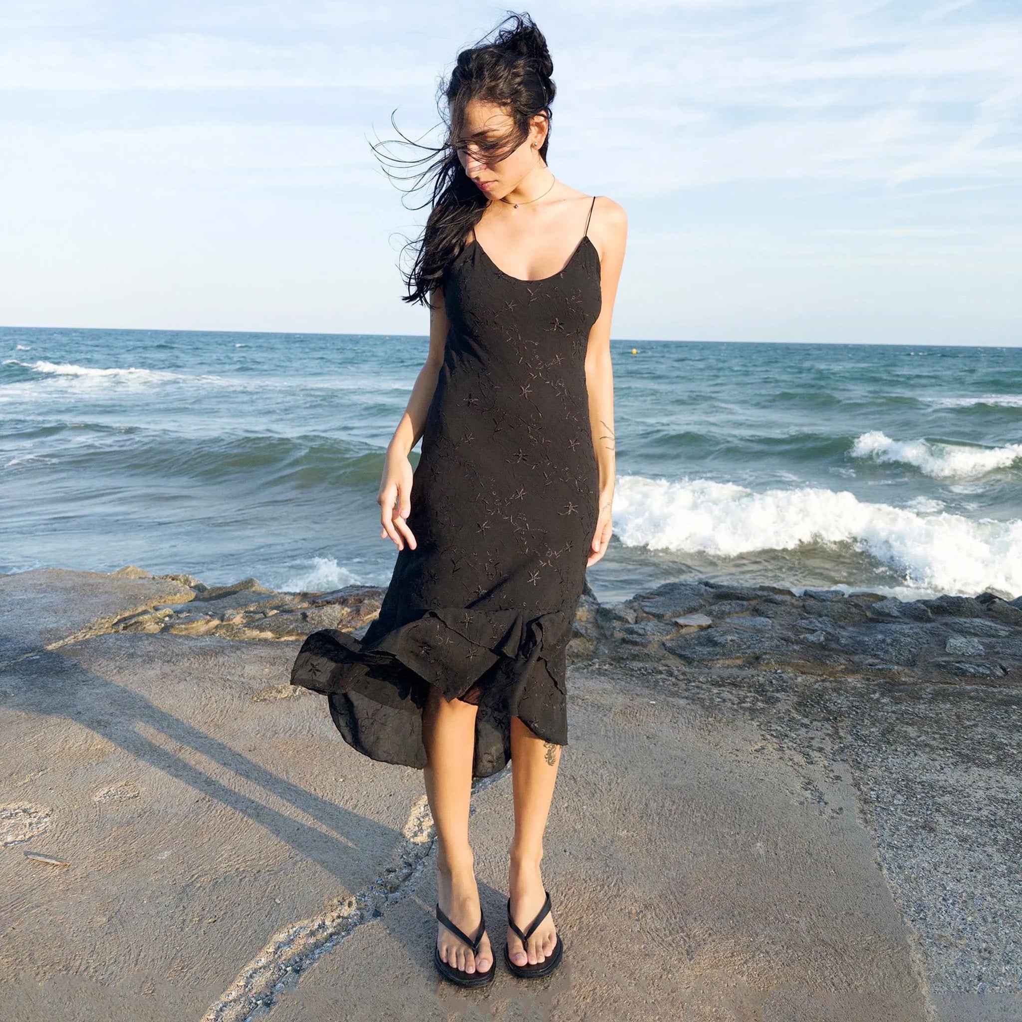 Vintage 2000s black midi dress with a slip silhouette, spaghetti straps, subtle floral embroidery, and a tiered ruffle hem that drapes asymmetrically. Lightweight and ideal for summer promenades, cocktail sunsets, or barefoot evenings. Made by a French brand. Excellent vintage condition.