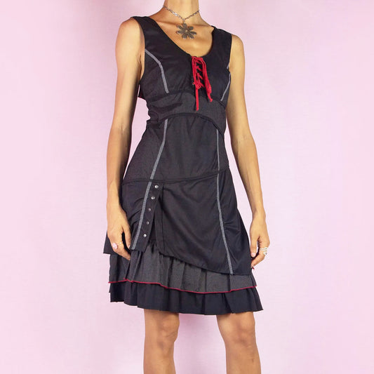 The Y2K Black Layered Lace-Up Dress is a vintage 2000s mini dress featuring a sleeveless design, scoop neckline, and stretchy fabric. It showcases a red lace-up detail at the bodice, contrasting stitching throughout, and a side zipper closure. The layered skirt, with its pinstripe and solid panels, button accents, and ruffle hem, adds a touch of whimsy. Perfect for night events and parties. Great vintage condition.
