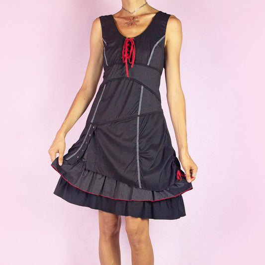 The Y2K Black Layered Lace-Up Dress is a vintage 2000s mini dress featuring a sleeveless design, scoop neckline, and stretchy fabric. It showcases a red lace-up detail at the bodice, contrasting stitching throughout, and a side zipper closure. The layered skirt, with its pinstripe and solid panels, button accents, and ruffle hem, adds a touch of whimsy. Perfect for night events and parties.
