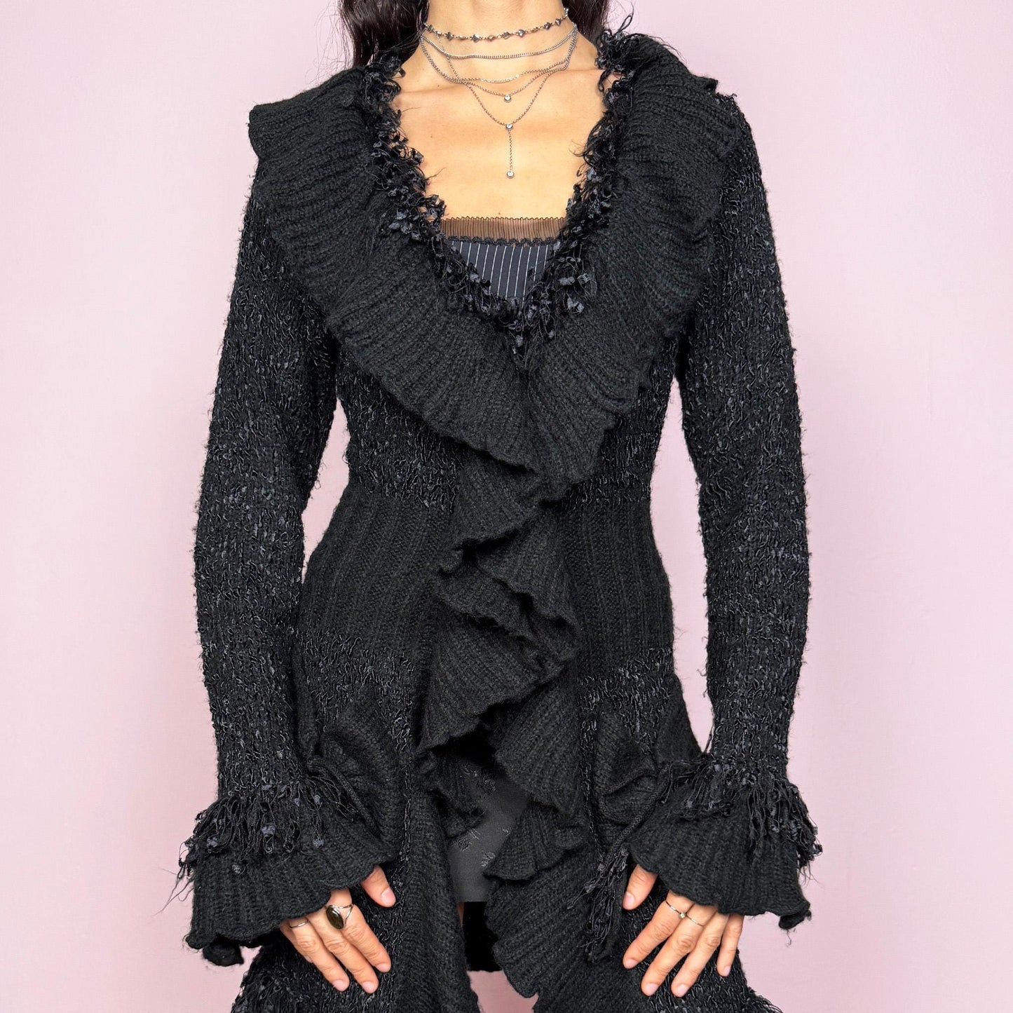 Witchy vintage 2000s black longline cardigan with dramatic ruffle neckline and dark romantic rococo style. Features bell sleeves with ruffle cuffs, two-button front closure, pockets, and asymmetric pointy hem. Whimsical fairycore silhouette, stretchy fabric for cozy winter layering. Made from polyester acrylic blend. Great vintage condition.