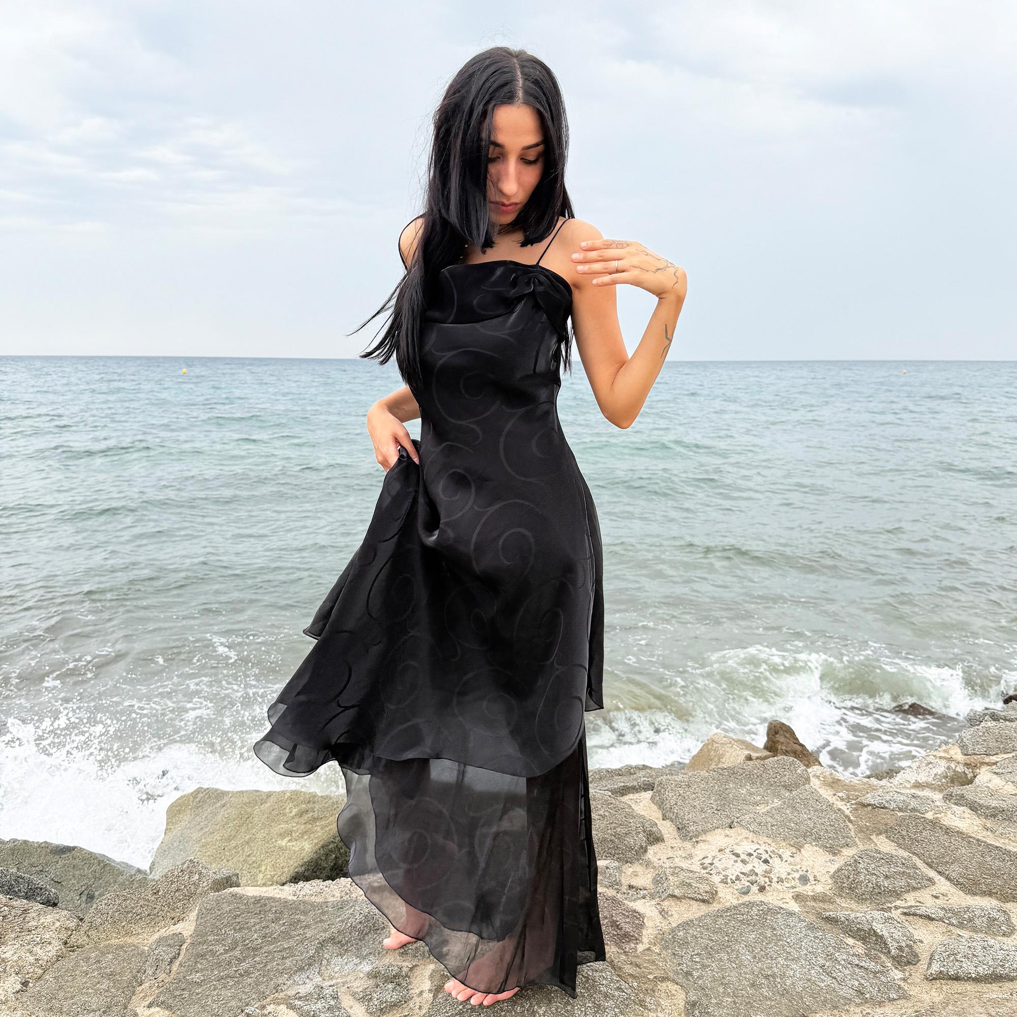 This vintage 2000s black organza-style layered slip midi dress is a French-made evening piece featuring a swirl pattern, folded neckline detail, delicate spaghetti straps, side zipper, and a flowing asymmetrical hem. It comes with a matching scarf, making it ideal for galas, proms, or dark, romantic nights. Crafted from a semi-sheer, non-stretch polyester blend. Excellent vintage condition.