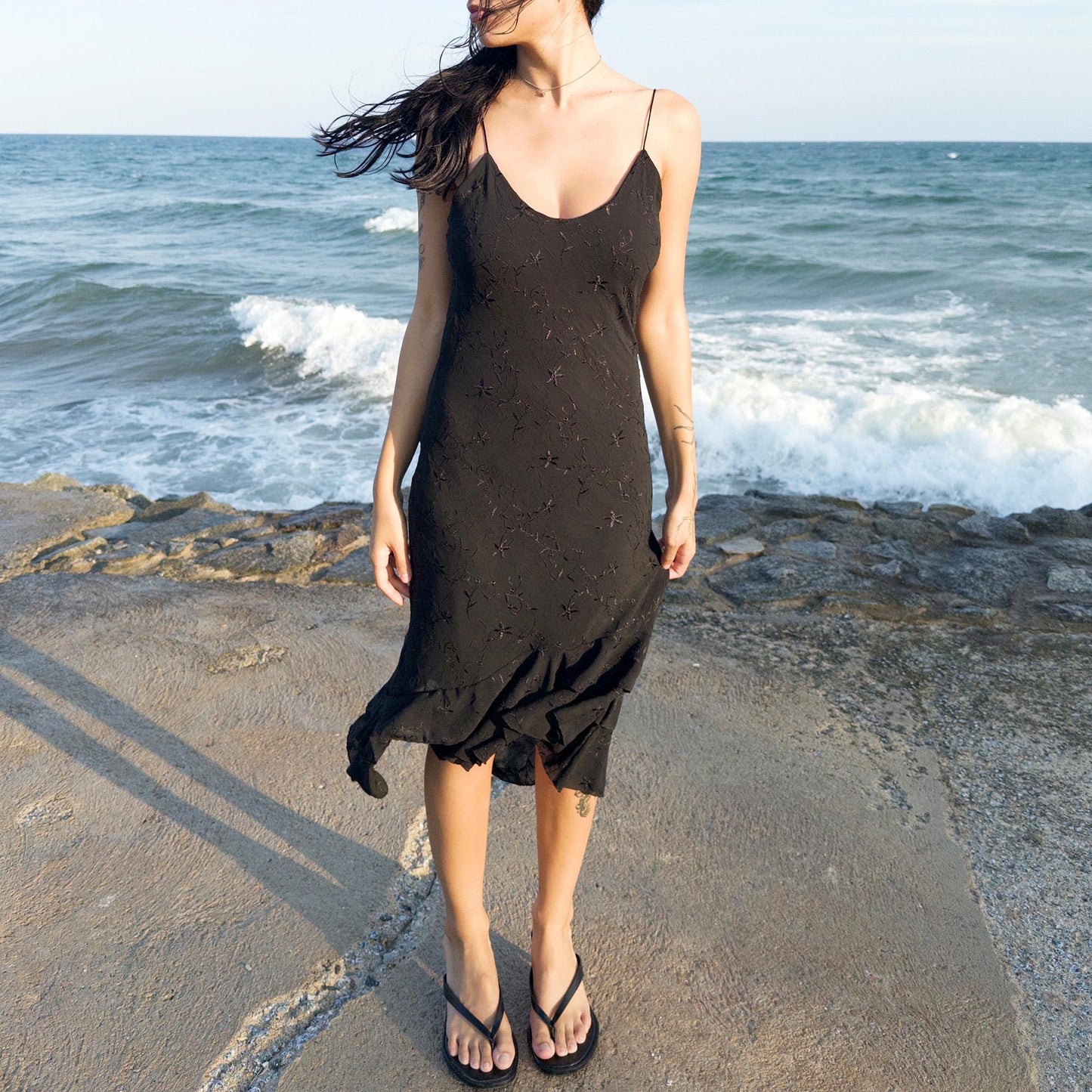 The vintage 2000s black midi dress is a French-made slip silhouette with delicate spaghetti straps and a tiered ruffle hem that drapes asymmetrically. Lightweight with subtle floral embroidery, it flows with ease—ideal for summer promenades, cocktail sunsets, or barefoot evenings. Its understated sensuality and effortless cut evoke a romantic calm. Excellent vintage condition.