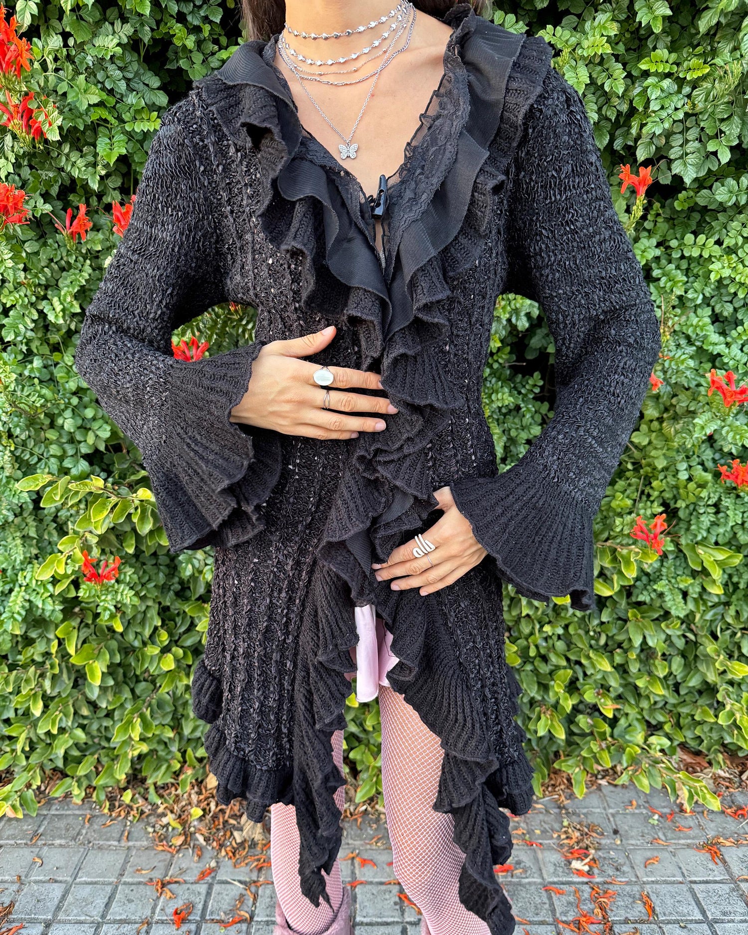 Vintage 2000s black longline cardigan featuring a dramatic ruffle neckline with lace details, bell-sleeve ruffle cuffs and a toggle front closure. The asymmetric pointy hem adds a whimsical, witchy silhouette, while the stretchy cotton–wool–polyamide blend creates a cozy, comfortable fit perfect for winter layering. A stunning fairy-goth statement knit jacket that blends elegance, softness and dark romantic style. Great vintage condition.