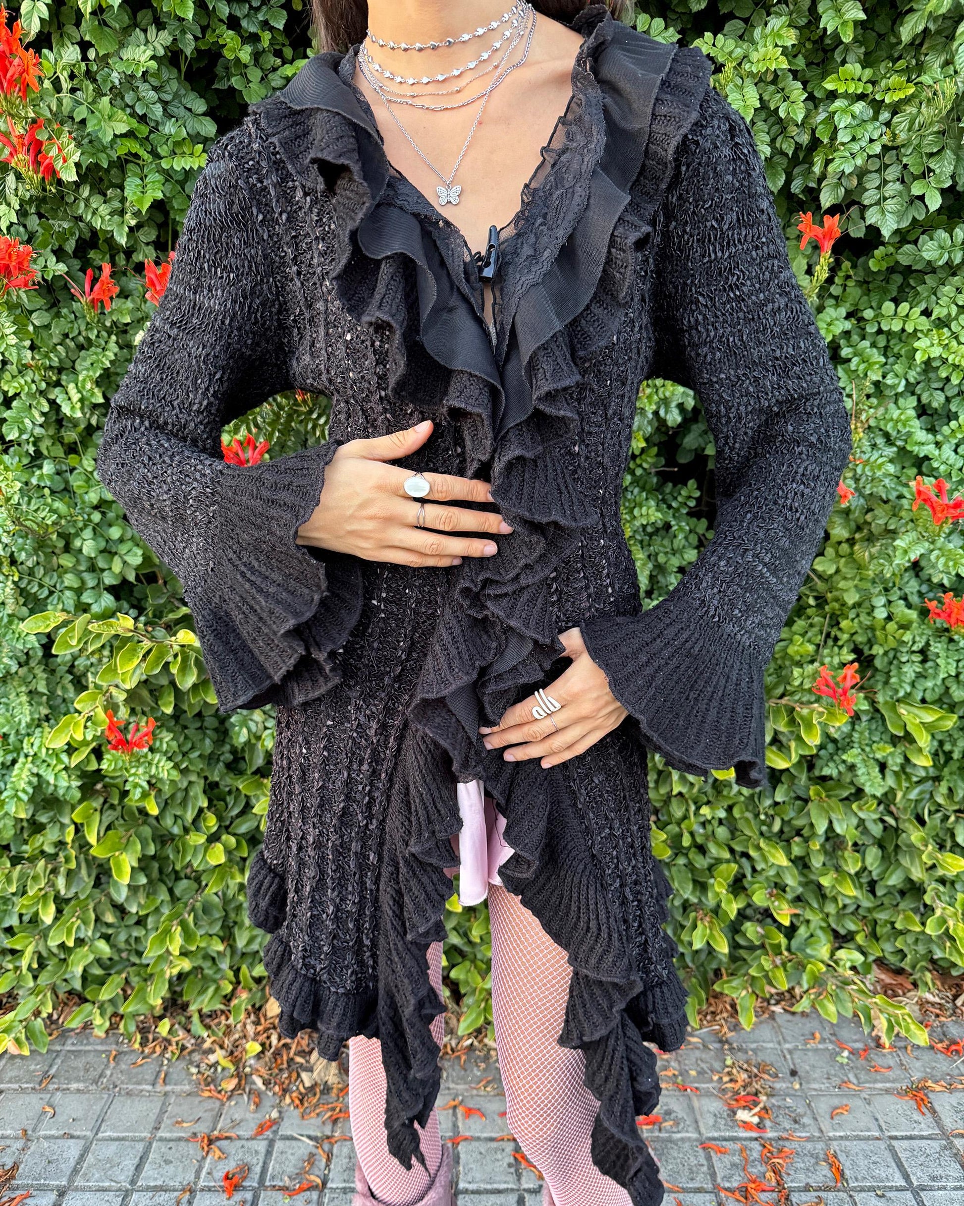 Vintage 2000s black longline cardigan featuring a dramatic ruffle neckline with lace details, bell-sleeve ruffle cuffs and a toggle front closure. The asymmetric pointy hem adds a whimsical, witchy silhouette, while the stretchy cotton–wool–polyamide blend creates a cozy, comfortable fit perfect for winter layering. A stunning fairy-goth statement knit jacket that blends elegance, softness and dark romantic style. Great vintage condition.