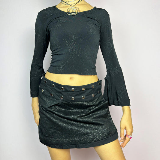 The Y2K Wrap Mini Black Skirt is a vintage 2000s piece featuring a snap-button closure on the waistband for an adjustable fit. Made from a thick fabric with a subtle floral pattern design, its wrap silhouette makes it perfect for casual outfits or festival looks. Excellent vintage condition.