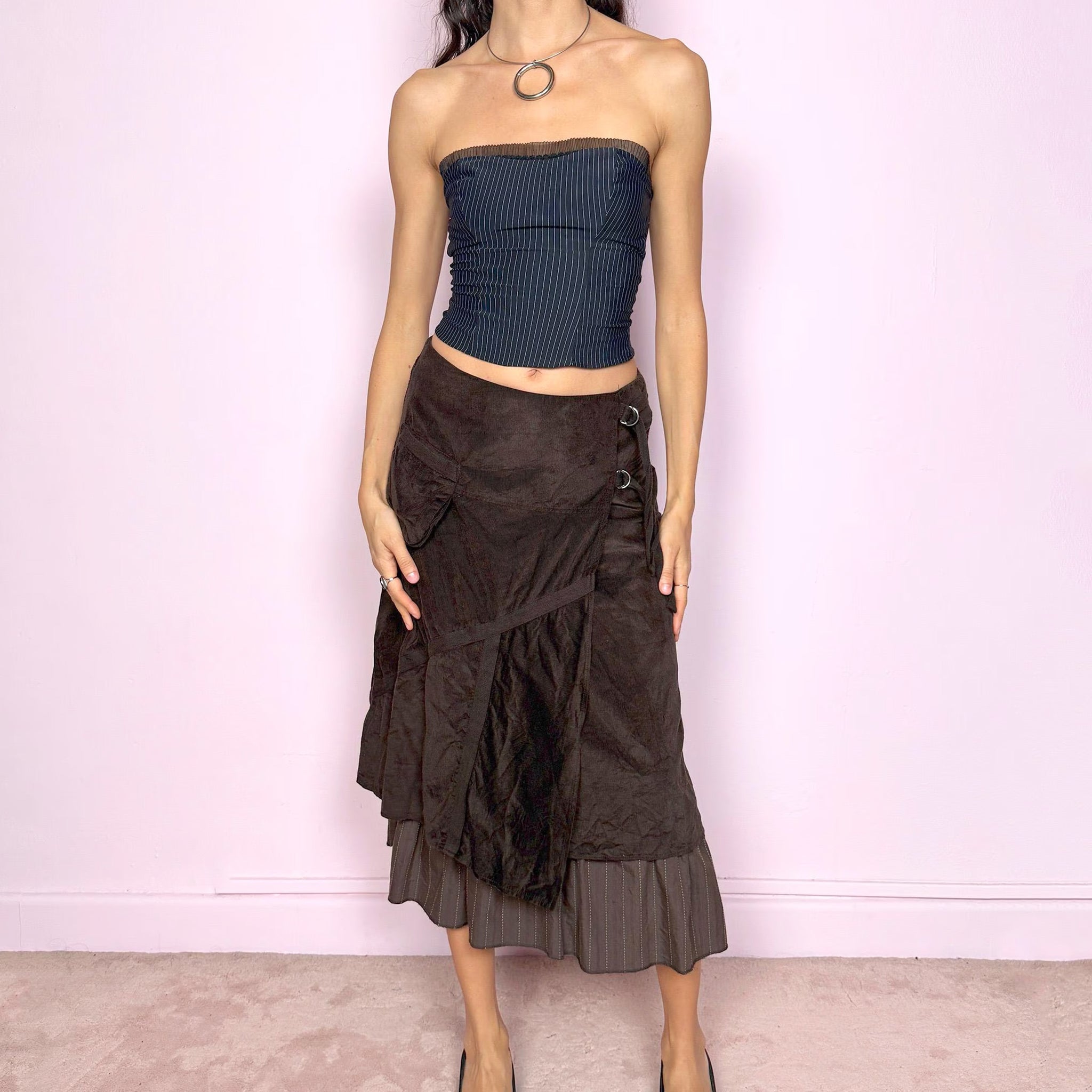 Vintage 2000s dark brown midi skirt with a layered wrap design and ruffled striped hem. Features decorative tie details, a back zipper closure, and a pocket for a deconstructed look. Ideal for earthy fairycore, bohemian, avant-garde, or cyber futuristic styling. Can be worn mid to low rise for size XXS–XS or high waisted for size S. Made from corduroy cotton and polyester. Great vintage condition.