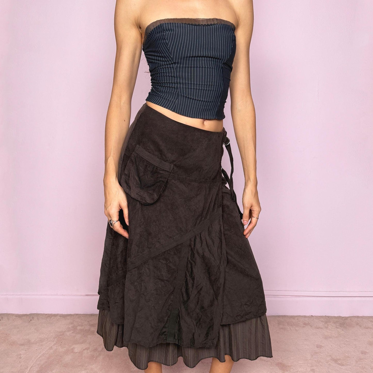 Vintage 2000s dark brown midi skirt with a layered wrap design and ruffled striped hem. Features decorative tie details, a back zipper closure, and a pocket for a deconstructed look. Ideal for earthy fairycore, bohemian, avant-garde, or cyber futuristic styling. Can be worn mid to low rise for size XXS–XS or high waisted for size S. Made from corduroy cotton and polyester. Great vintage condition.