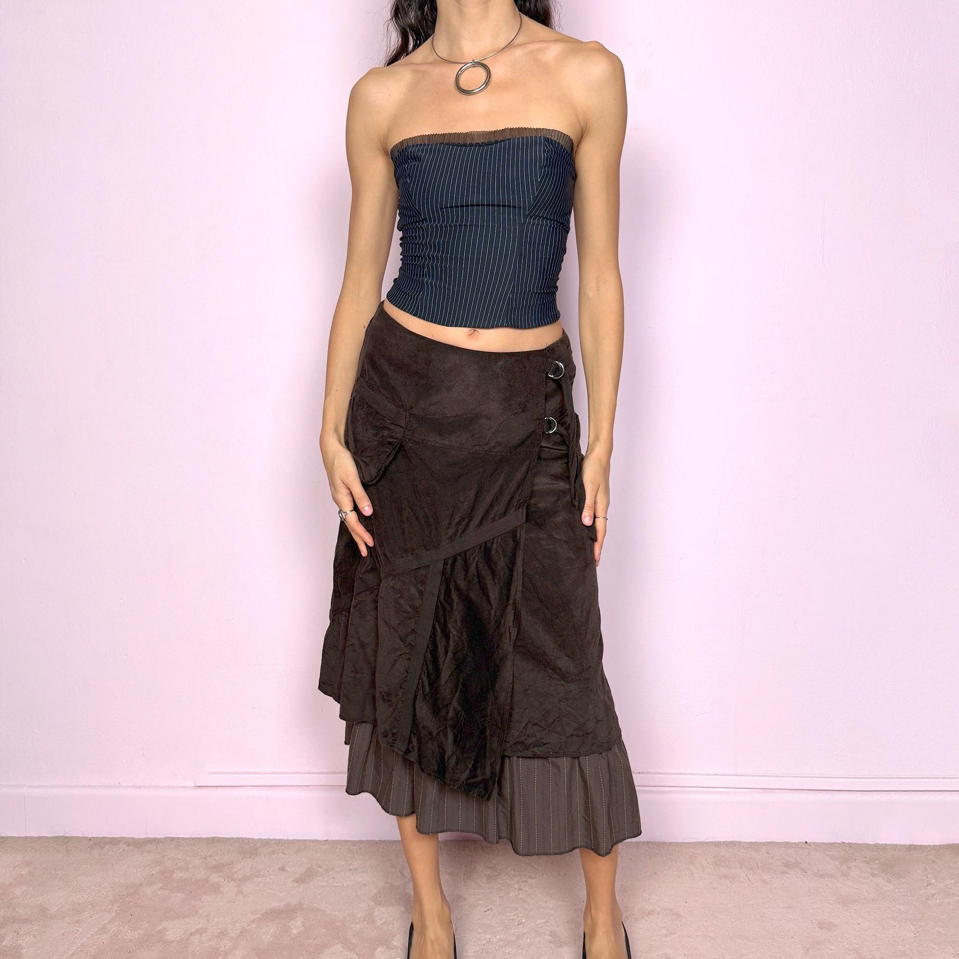 Vintage 2000s dark brown midi skirt with a layered wrap design and ruffled striped hem. Features decorative tie details, a back zipper closure, and a pocket for a deconstructed look. Ideal for earthy fairycore, bohemian, avant-garde, or cyber futuristic styling. Can be worn mid to low rise for size XXS–XS or high waisted for size S. Made from corduroy cotton and polyester. Great vintage condition.
