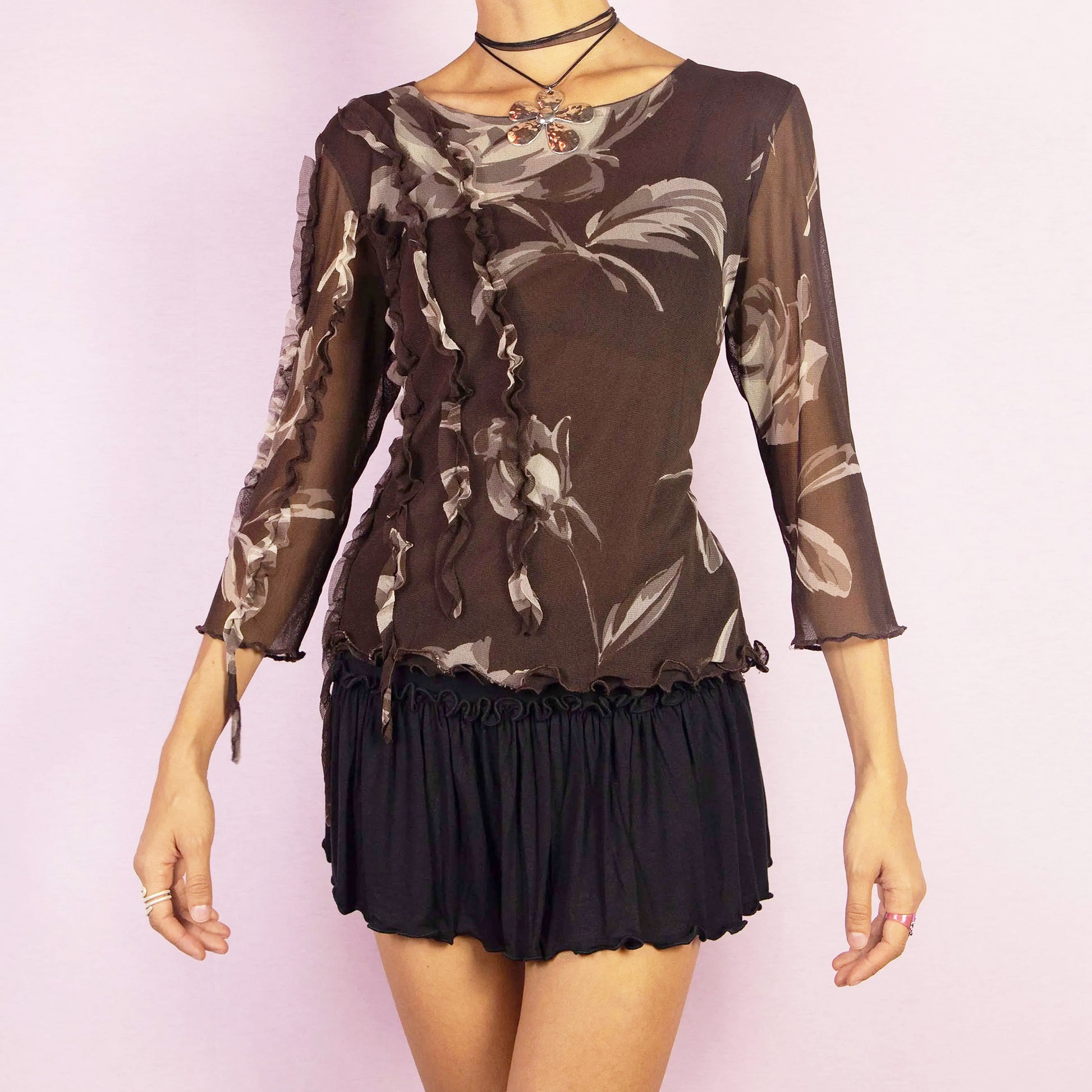 The Y2K Brown Frill Mesh Top is a vintage 2000s blouse featuring a semi-sheer fabric with a floral print in earthy brown tones. The top is detailed with whimsical ruffle accents along the front and sleeves, an asymmetric design, and three-quarter sleeves. Perfect for casual or evening looks.