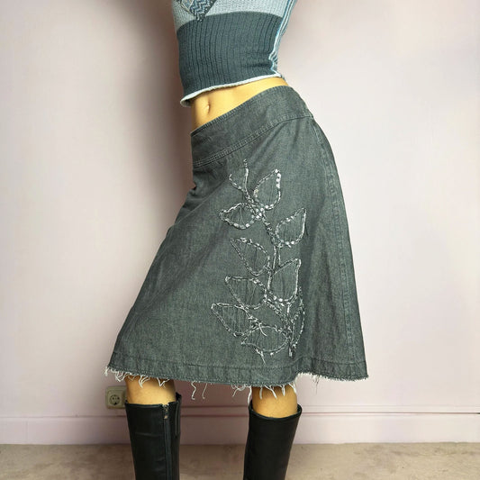 The Y2K Denim Gray Midi Skirt is a vintage 2000s piece featuring a faded gray wash, raw hem, and a unique floral sequin embroidery design. Crafted from structured denim, this skirt has an A-line silhouette that drapes effortlessly. Designed with a back zipper closure, the unfinished hem adds a deconstructed, edgy touch, making it perfect for casual outfits.