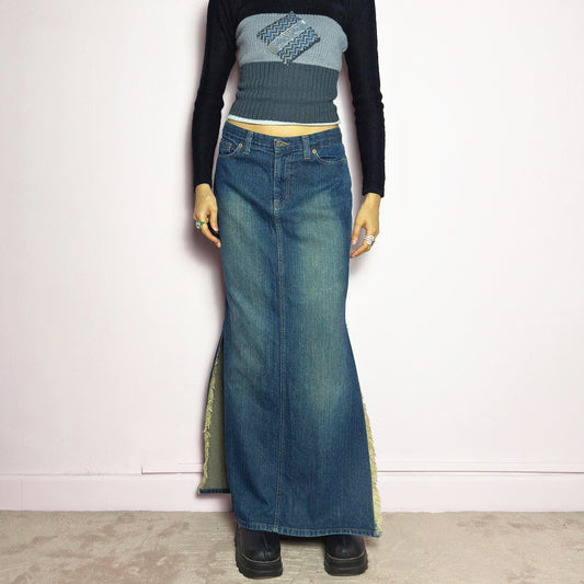 The Y2K Denim Side Slit Maxi Skirt is a vintage 2000s statement piece featuring a structured mid-rise fit and a sleek, elongated silhouette. Designed with frayed side slit details, a button and zipper closure, and classic five-pocket styling, this skirt is perfect for casual streetwear looks. Excellent vintage condition.