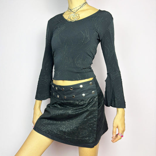 The Y2K Wrap Mini Black Skirt is a vintage 2000s piece featuring a snap-button closure on the waistband for an adjustable fit. Made from a thick fabric with a subtle floral pattern design, its wrap silhouette makes it perfect for casual outfits or festival looks.