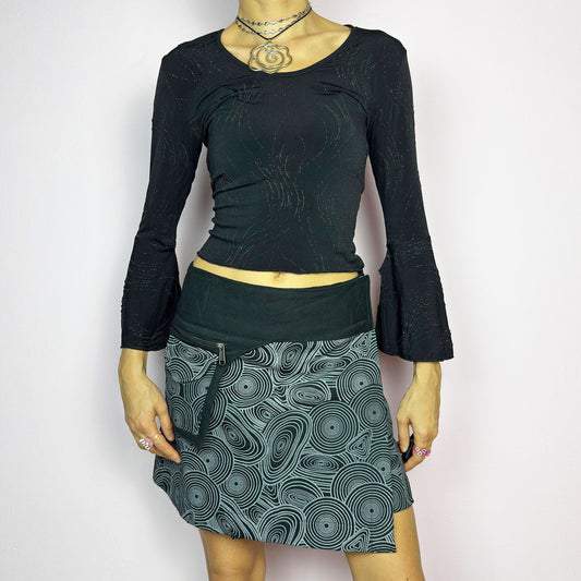 The Y2K Wrap Black Mini Skirt is a vintage 2000s piece designed with a Velcro wrap closure for an adjustable fit. Made from a thick fabric, it features a bold geometric swirl print and a utility pocket with a zipper closure. Perfect for casual and festival looks.