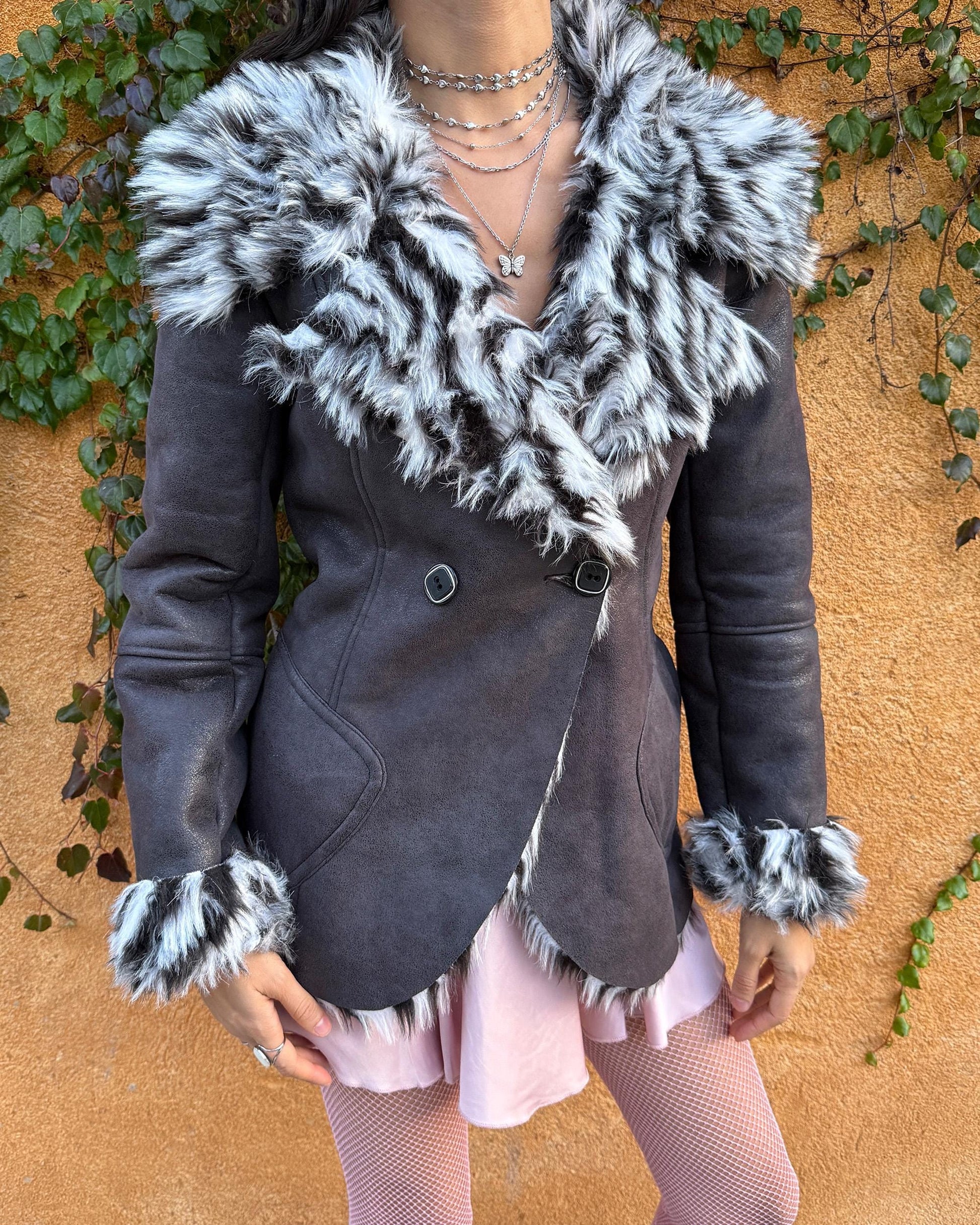 Vintage 2000s dark gray, almost black faux leather Penny Lane jacket with bold faux fur lining at the collar, cuffs, and hem in a black-and-white zebra-inspired pattern. Features a double row of buttons and side pockets. The dramatic fur trim gives it an iconic Y2K glam look, perfect for party nights or bold winter styling. Warm, comfortable, and eye-catching with strong 2000s attitude. Made from a polyester acrylic blend. Excellent vintage condition.