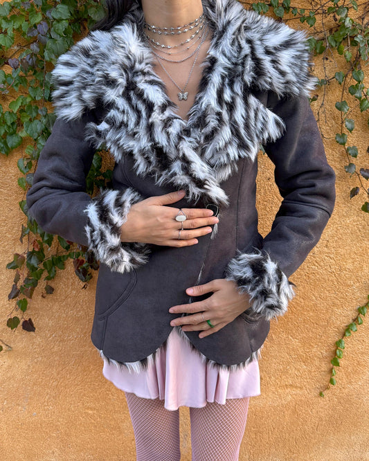 Vintage 2000s dark gray, almost black faux leather Penny Lane jacket with bold faux fur lining at the collar, cuffs, and hem in a black-and-white zebra-inspired pattern. Features a double row of buttons and side pockets. The dramatic fur trim gives it an iconic Y2K glam look, perfect for party nights or bold winter styling. Warm, comfortable, and eye-catching with strong 2000s attitude. Made from a polyester acrylic blend. Excellent vintage condition.
