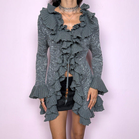 This vintage 2000s gray longline cardigan features cascading ruffles, a tie-front closure, and dramatic bell sleeves. Its sculptural knit design blends romance and whimsy, perfect for fairycore or Y2K-inspired layering. The mohair-acrylic blend adds a soft shimmer and cozy texture, making it ideal for autumn evenings or winter parties. A rare statement knit that elevates any outfit from casual to editorial. Made from mohair acrylic blend. Excellent vintage condition.