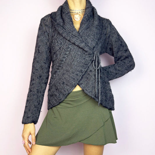 The Y2K Gray Wrap Tie Cardigan is a cozy vintage 2000s piece. This dark gray cardigan features a chunky knit design with intricate cable-knit detailing and a wrap-front closure that ties at the side for an adjustable fit. Perfect for layering during autumn and winter. Great vintage condition.