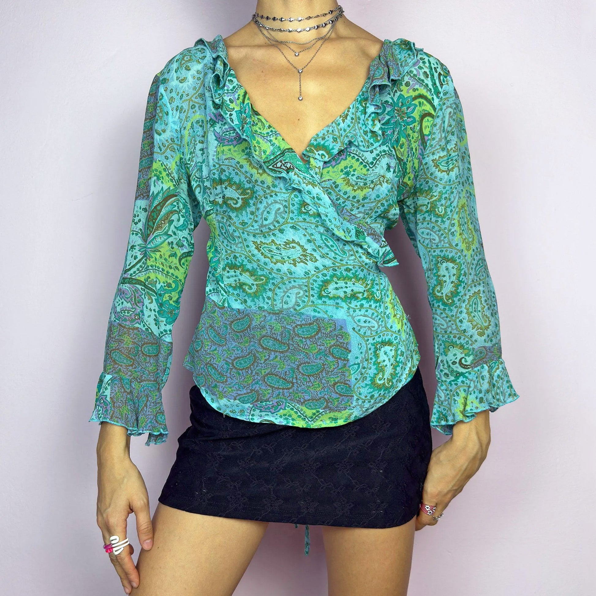 The Y2K Ruffle Wrap Tie Blouse is a vintage 2000s vibrant and playful summer statement piece, showcasing a lively green and blue paisley print with a semi-sheer finish. Featuring a flattering wrap-front design with adjustable ties at the back, ruffled detailing along the neckline and cuffs adds a breezy, feminine touch. This lightweight blouse is perfect for warm-weather outfits.