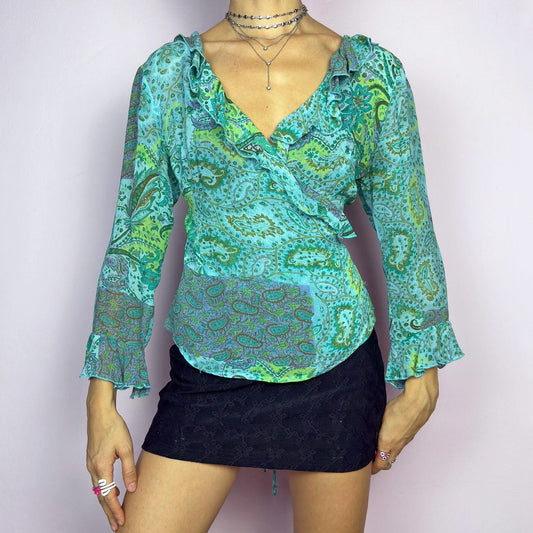 The Y2K Ruffle Wrap Tie Blouse is a vintage 2000s vibrant and playful summer statement piece, showcasing a lively green and blue paisley print with a semi-sheer finish. Featuring a flattering wrap-front design with adjustable ties at the back, ruffled detailing along the neckline and cuffs adds a breezy, feminine touch. This lightweight blouse is perfect for warm-weather outfits.