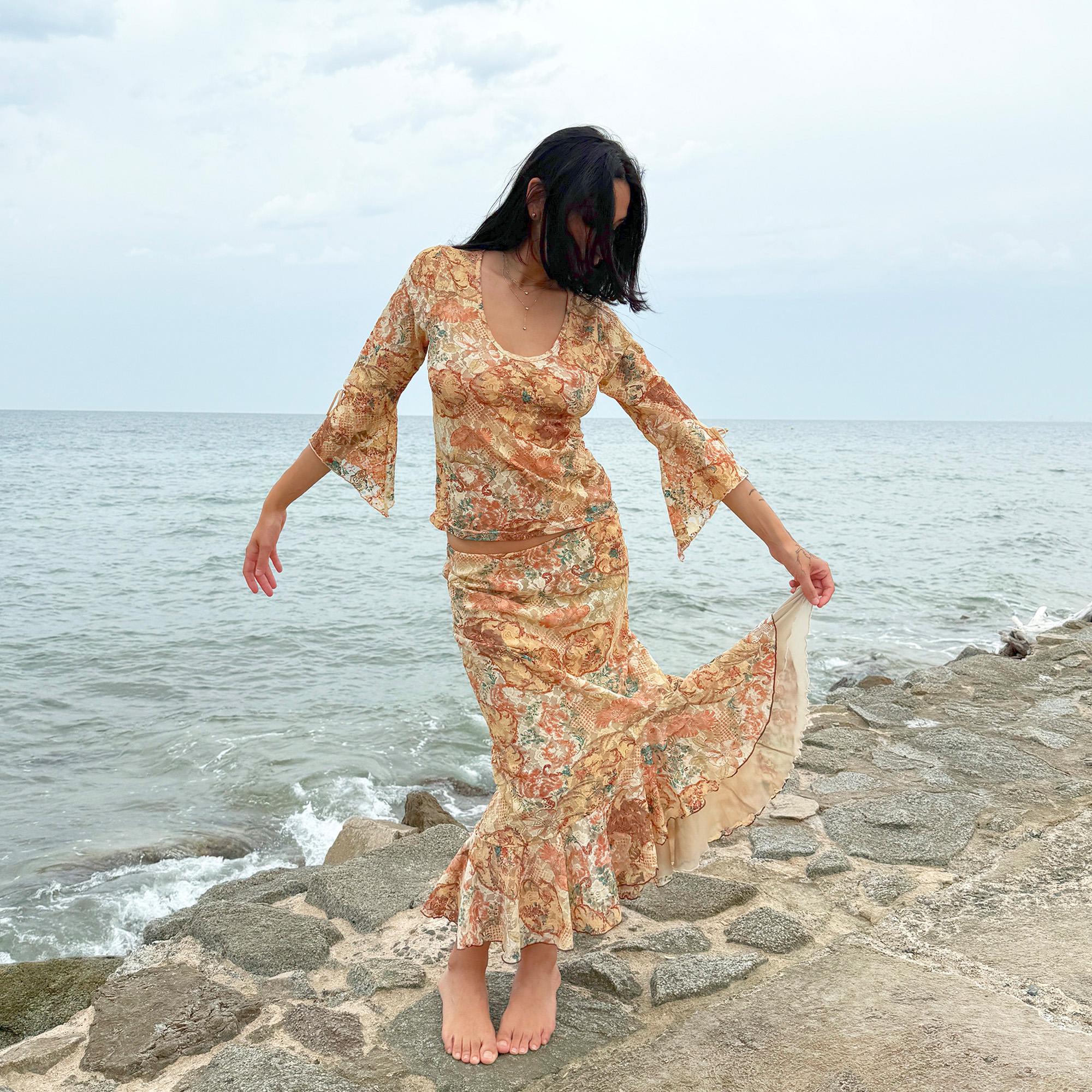 This vintage 2000s two-piece set features a cream-beige floral lace mesh with brown and blue accents. The asymmetric midi skirt has a flowing ruffle hem, paired with a matching semi sheer top with a scoop neckline and three-quarter tie bell sleeves. A whimsical spring-summer archival statement, ideal for parties, festivals, and seaside escapes. Made in Spain from polyester stretch mesh. Excellent vintage condition.
