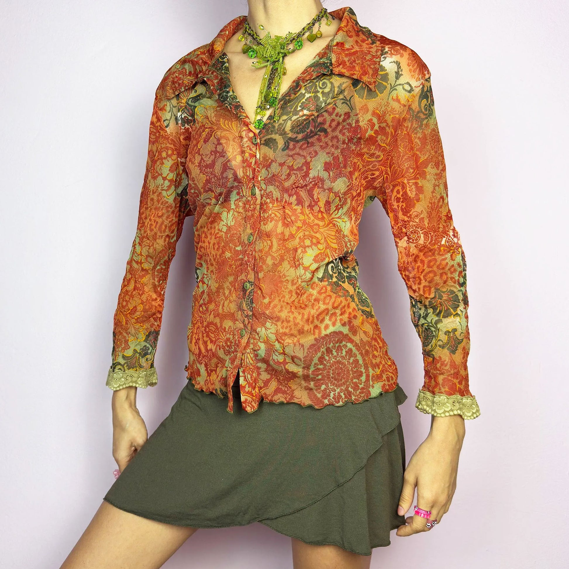 The Y2K Baroque Print Mesh Blouse is a vintage 2000s piece featuring a semi-sheer mesh fabric with a rich baroque-inspired floral print in warm tones. Designed with a fitted silhouette, button-up front, delicate lace trim on the cuffs and three quarter sleeves, the lightweight and stretchy fabric make it ideal for summer bohemian-inspired looks.
