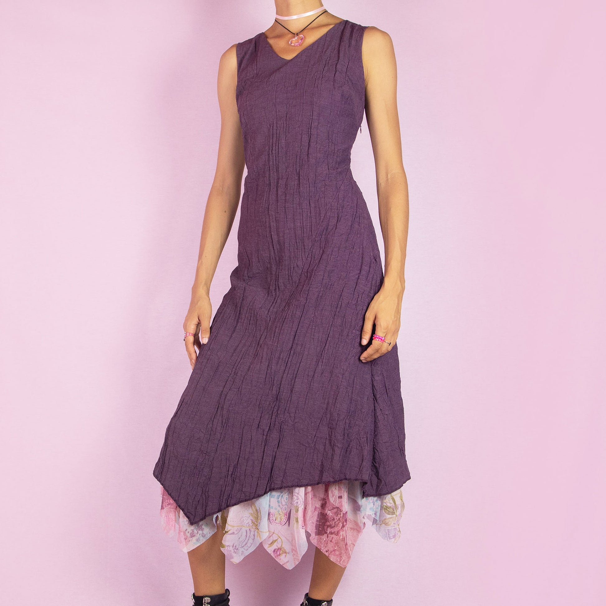 The Y2K Purple Sleeveless Midi Dress is a vintage 2000s piece featuring a crinkled, textured fabric, a V-neckline, and a side zipper closure. The dress also has a layered asymmetric hemline with an abstract floral print mesh sheer underlayer peeking out. Perfect for summer outings and elegant parties.
