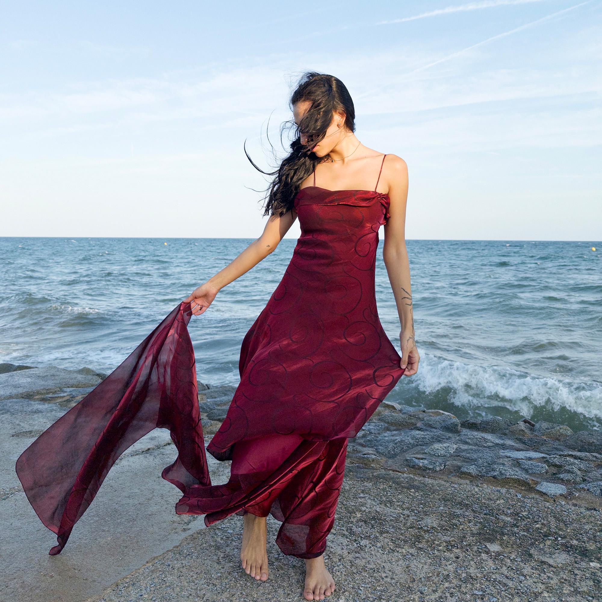 The vintage 2000s red organza-style slip midi dress is a French-made evening piece with layered maroon tones and tonal swirl pattern. With delicate spaghetti straps, side zip, and a flowing asymmetrical hem, it includes a matching scarf. Ideal for galas, proms, or romantic nights under open skies. Crafted from a semi-sheer, non-stretch fabric. Excellent vintage condition.