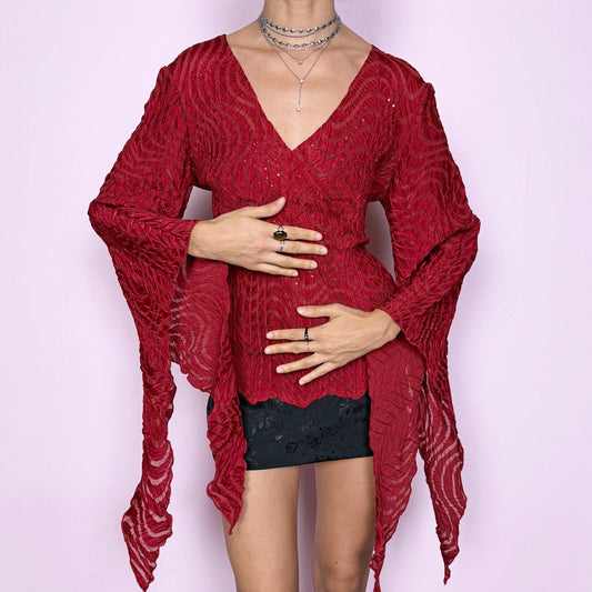 This romantic glam vintage 2000s red blouse features a sheer burnout textured pattern with crinkled wavy detailing and subtle sequins. Designed with a wrap-style silhouette, three-quarter slit dramatic bell sleeves, and an asymmetric hem, it offers a sculpted yet fluid fit ideal for artistic parties, nights out, or editorial avant-garde styling. Made from stretchy polyester. Great vintage condition.