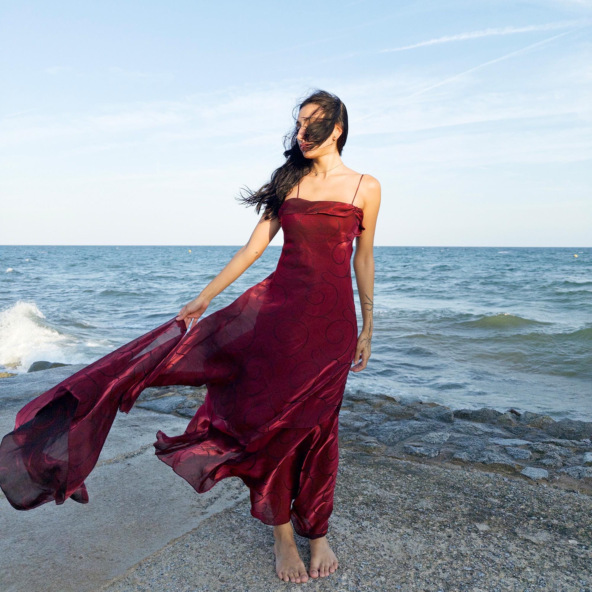 The vintage 2000s red organza-style slip midi dress is a French-made evening piece with layered maroon tones and tonal swirl pattern. With delicate spaghetti straps, side zip, and a flowing asymmetrical hem, it includes a matching scarf. Ideal for galas, proms, or romantic nights under open skies. Crafted from a semi-sheer, non-stretch fabric. Excellent vintage condition.