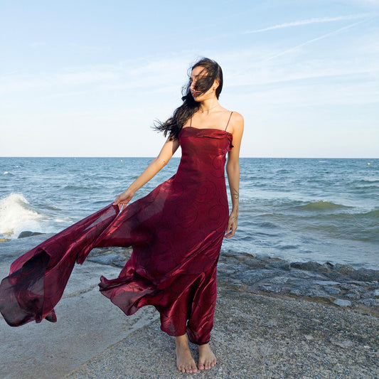 The vintage 2000s red organza-style slip midi dress is a French-made evening piece with layered maroon tones and tonal swirl pattern. With delicate spaghetti straps, side zip, and a flowing asymmetrical hem, it includes a matching scarf. Ideal for galas, proms, or romantic nights under open skies. Crafted from a semi-sheer, non-stretch fabric. Excellent vintage condition.