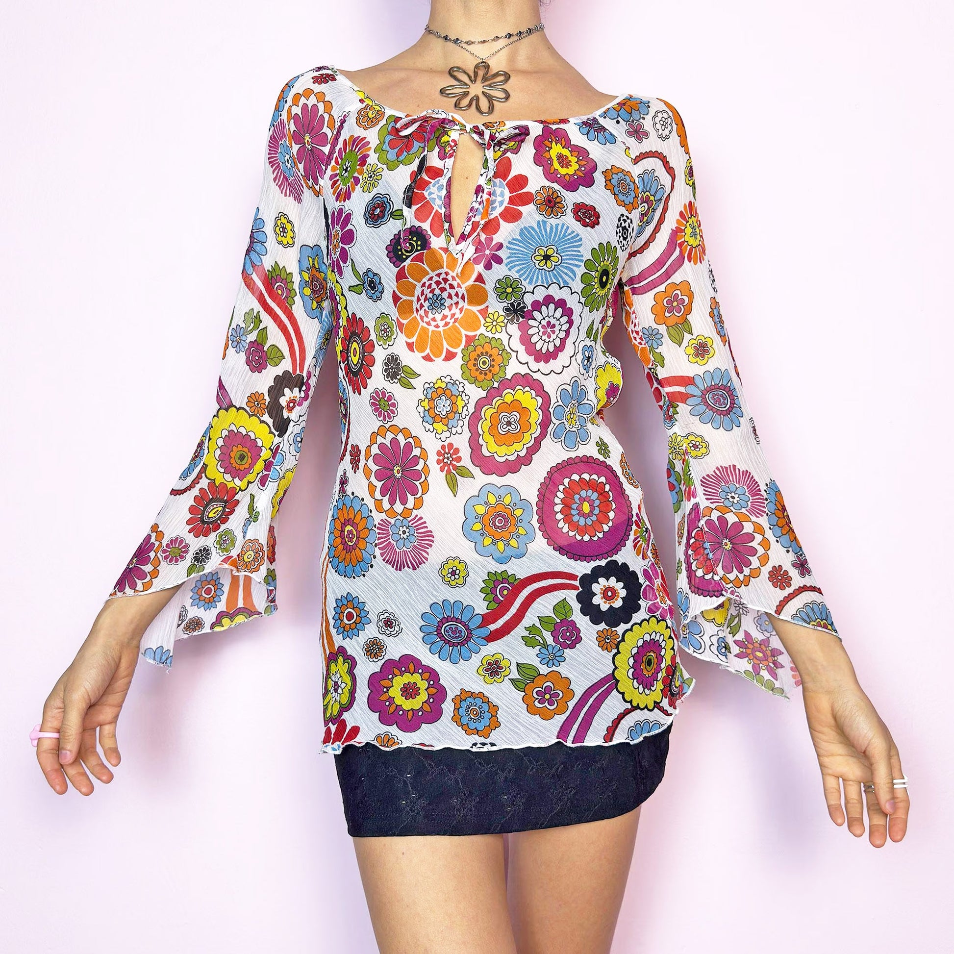The Y2K White Floral Tie Front Top is a vintage 2000s piece bursting with color and retro charm. Features a vibrant multicolor 60s-inspired flower print on lightweight, slightly sheer pleated fabric. Designed with a flattering scoop neckline, keyhole front with tie closure, and flared bell sleeves. Stretchy and breathable – perfect for summer, festivals, or hippie styles. Great vintage condition.