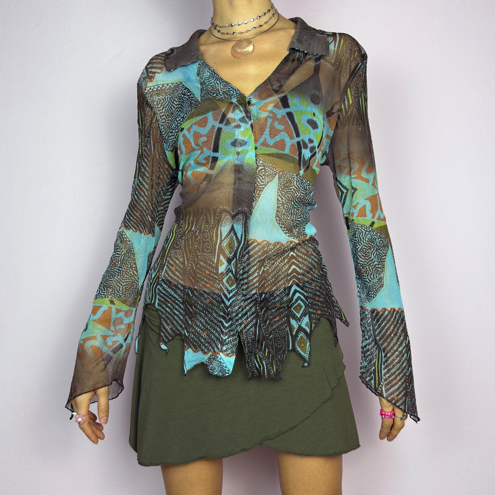 The Y2K Abstract Mesh Blouse is a vintage 2000s edgy and whimsical piece featuring a semi-sheer mesh fabric with bold, abstract patterns in earthy and blue tones and a button-front closure. The long flared sleeves. adult world shop
