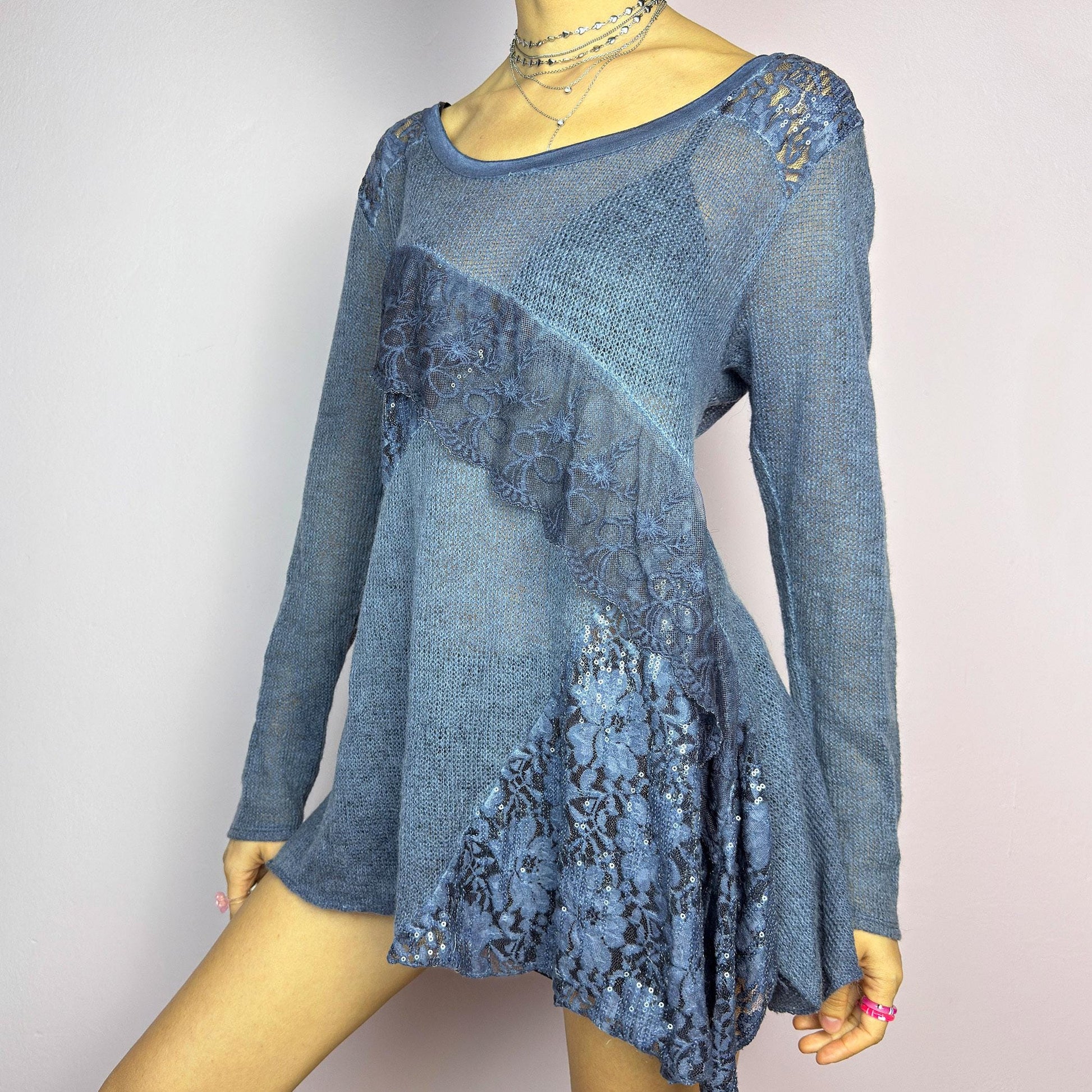 The Y2K Blue Lace Asymmetric Sweater is a vintage 2000s piece featuring a semi-sheer knit design with delicate lace and sequin paneling in an asymmetrical pattern. Designed with a scoop neckline, long sleeves, and a flowy silhouette, this sweater tunic adds a dreamy, romantic touch to any outfit. Perfect for layering in autumn and winter. adult world shop