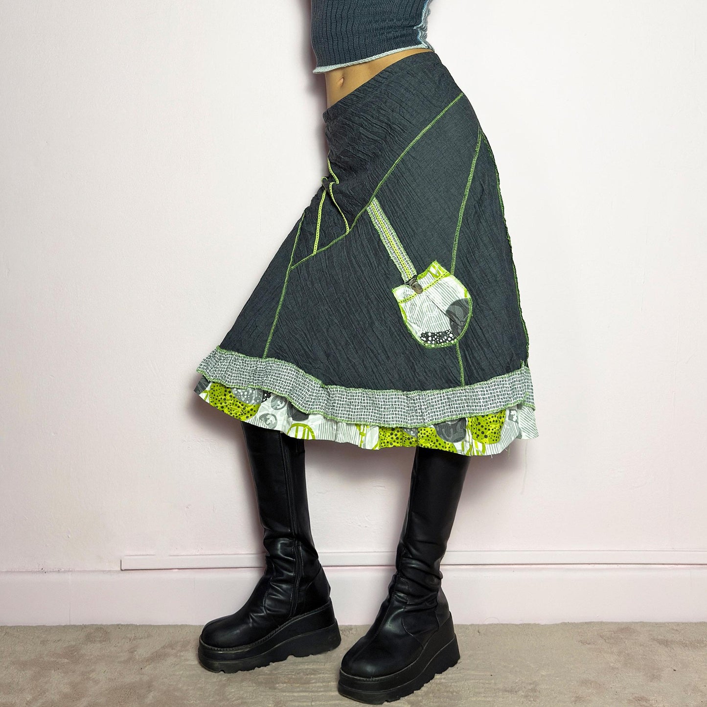 The Y2K Gray Patchwork Midi Skirt is a unique vintage 2000s piece featuring an eclectic mix of textures and contrasting neon green stitch details. Designed with a layered hem, ruffle trim, and a statement patch pocket with buckle detail, this skirt embodies a deconstructed boho aesthetic. The lightweight, crinkled fabric drapes effortlessly, while the elastic waistband ensures a comfortable fit, making it a perfect choice for bold, artsy outfits. Made in France. adult world shop