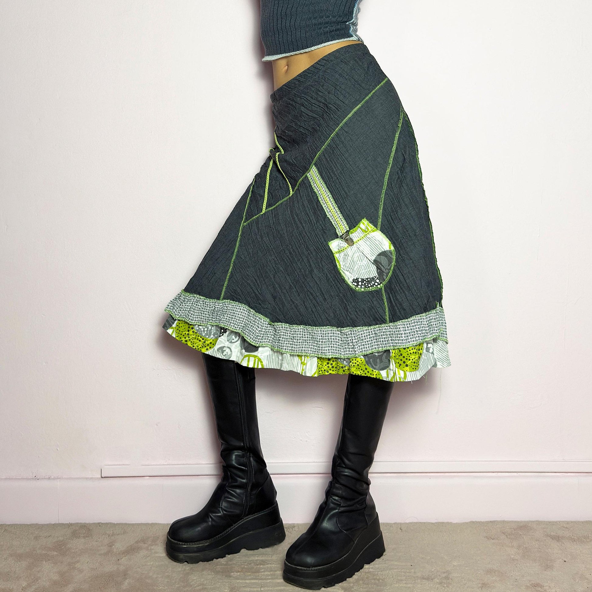 The Y2K Gray Patchwork Midi Skirt is a unique vintage 2000s piece featuring an eclectic mix of textures and contrasting neon green stitch details. Designed with a layered hem, ruffle trim, and a statement patch pocket with buckle detail, this skirt embodies a deconstructed boho aesthetic. The lightweight, crinkled fabric drapes effortlessly, while the elastic waistband ensures a comfortable fit, making it a perfect choice for bold, artsy outfits. Made in France. adult world shop
