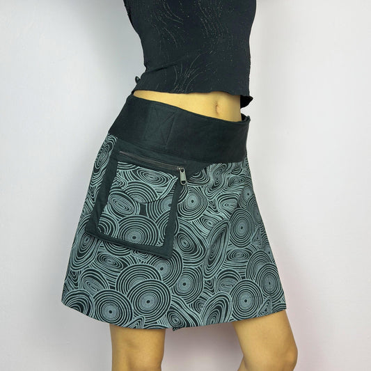The Y2K Wrap Black Mini Skirt is a vintage 2000s piece designed with a Velcro wrap closure for an adjustable fit. Made from a thick fabric, it features a bold geometric swirl print and a utility pocket with a zipper closure. adult world shop