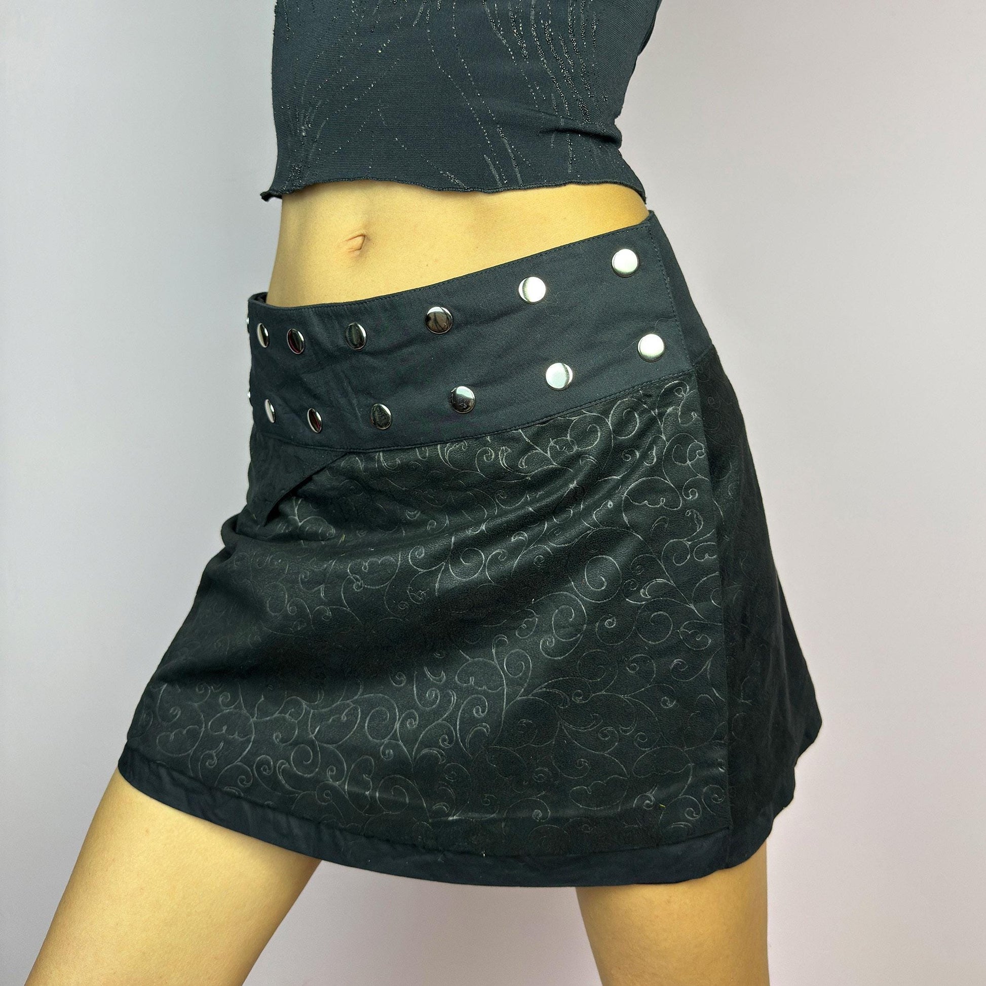 The Y2K Wrap Mini Black Skirt is a vintage 2000s piece featuring a snap-button closure on the waistband for an adjustable fit. Made from a thick fabric with a subtle floral pattern design, its wrap silhouette. adult world shop