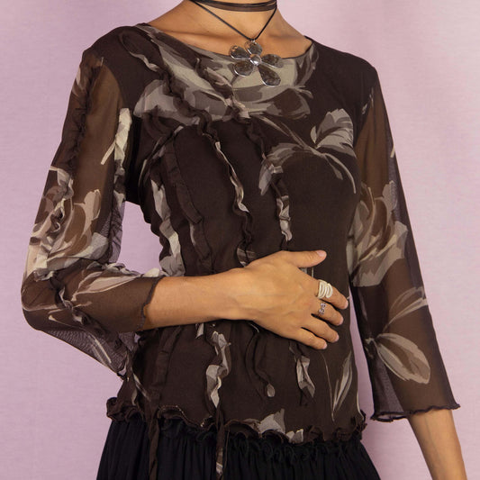 The Y2K Brown Frill Mesh Top is a vintage 2000s blouse featuring a semi-sheer fabric with a floral print in earthy brown tones. The top is detailed with whimsical ruffle accents along the front and sleeves, an asymmetric design