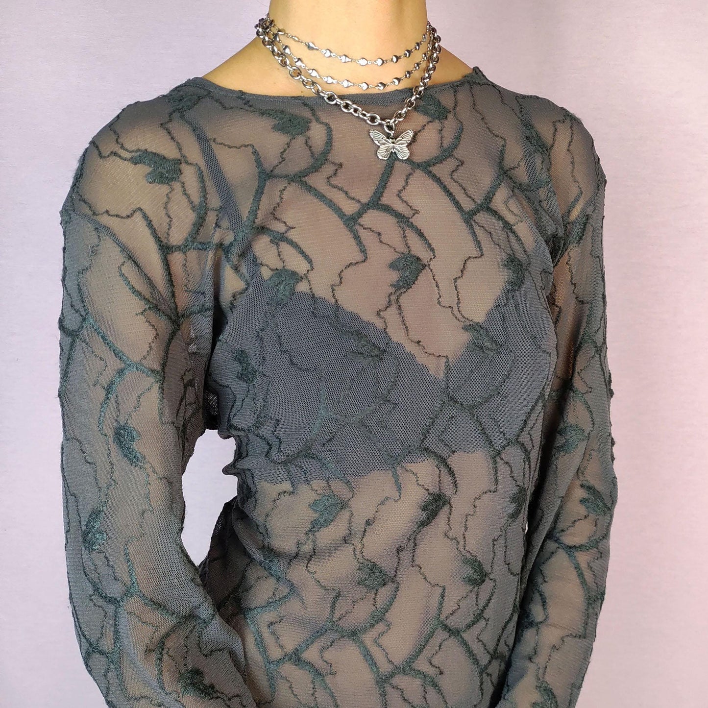The Y2K Gray Sheer Mesh Top is a vintage 2000s piece featuring a semi-sheer design with intricate textured patterns. Crafted from soft, lightweight mesh fabric, it has a slim-fit silhouette with long sleeves, making it perfect for layering