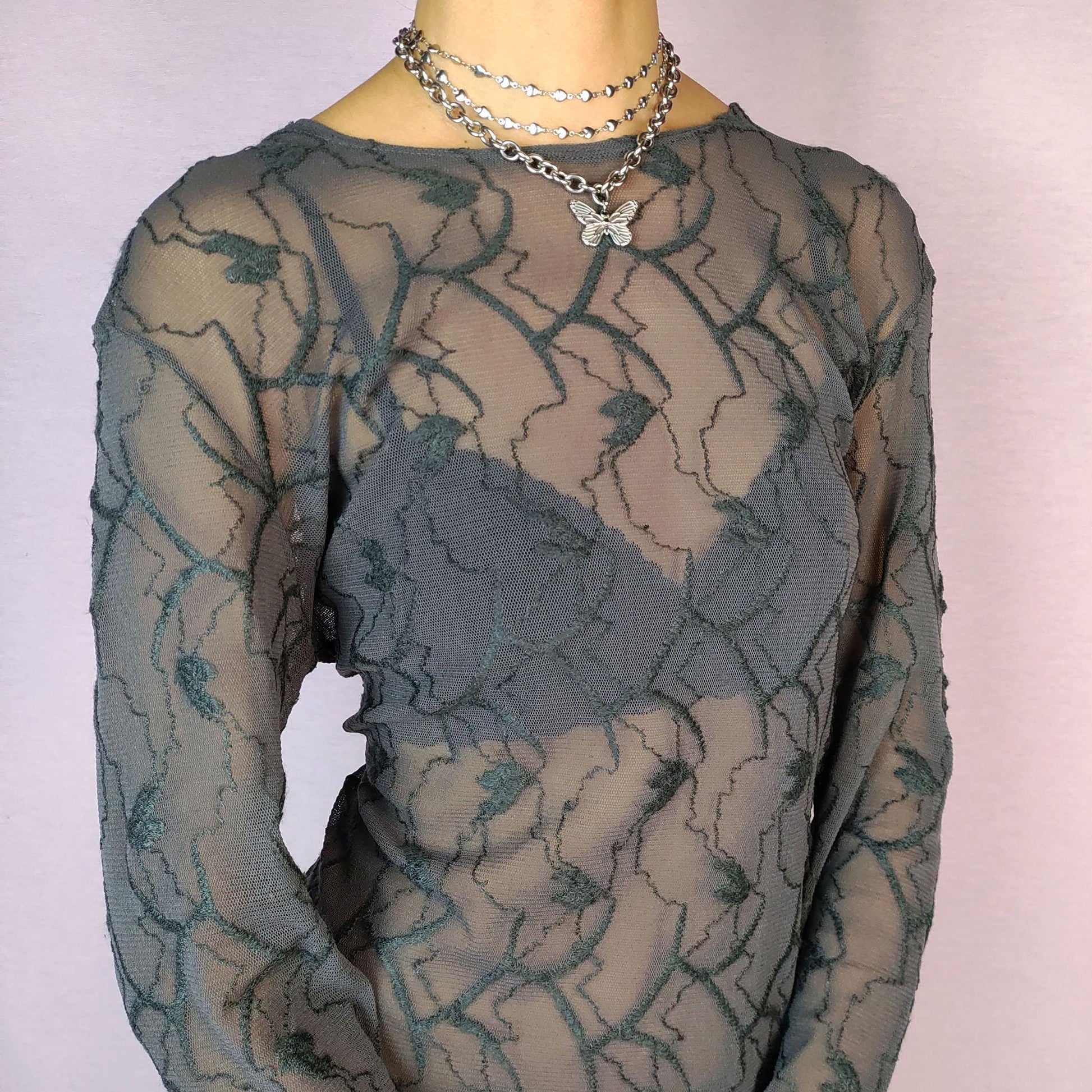 The Y2K Gray Sheer Mesh Top is a vintage 2000s piece featuring a semi-sheer design with intricate textured patterns. Crafted from soft, lightweight mesh fabric, it has a slim-fit silhouette with long sleeves, making it perfect for layering