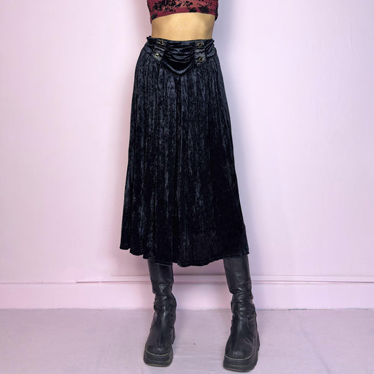 The Vintage 90s Black Velvet Midi Skirt is crafted from soft velvet and features an elastic waistband detail with button accents and a flowing A-line silhouette. Ideal for night parties and embodies a whimsical goth aesthetic. Adult World Shop