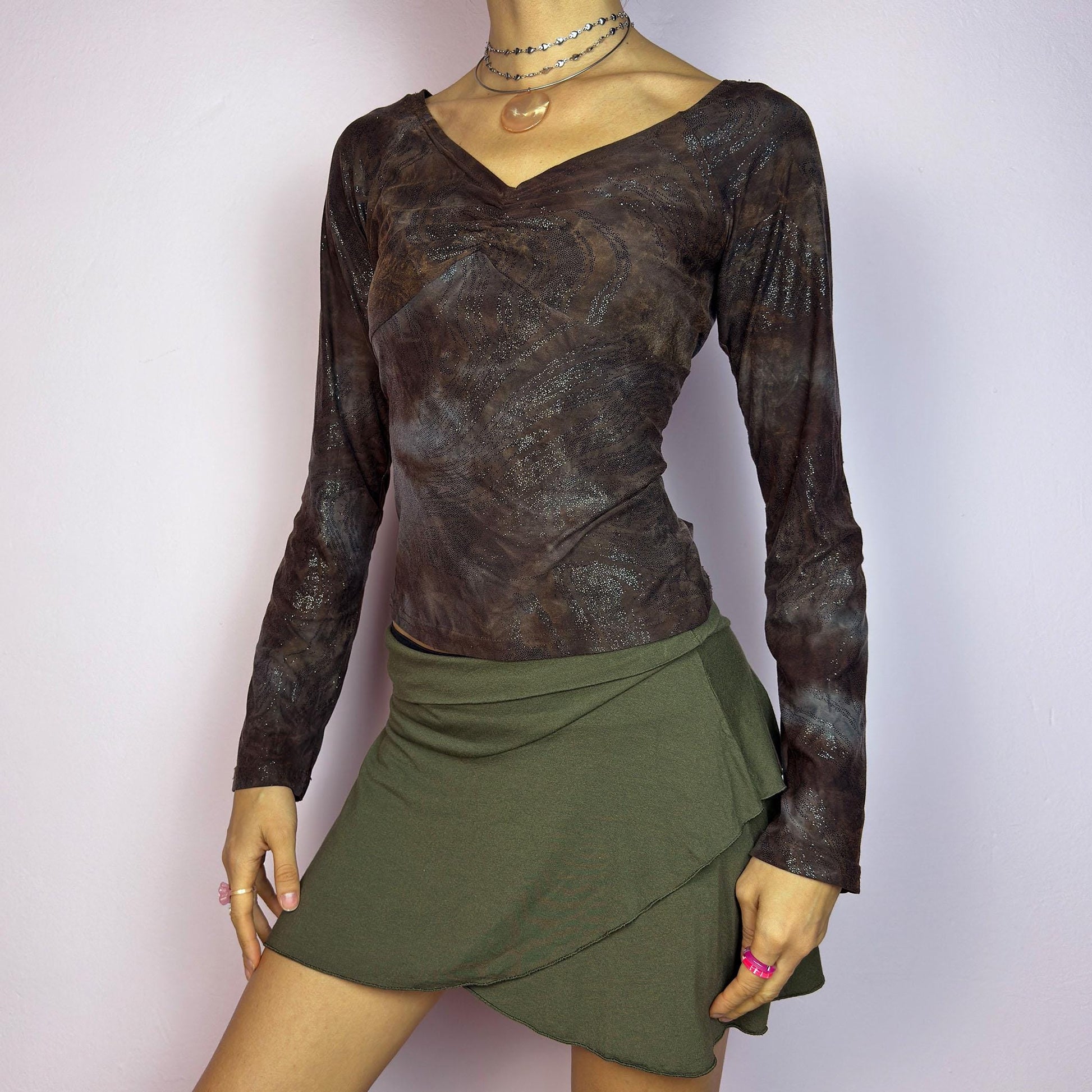 The Y2K Brown Earthy Shimmer Top is a vintage 2000s fitted and stretchy long-sleeve blouse with an earthy, abstract pattern featuring a ruched detail at the bust and subtle shimmering accents throughout. Perfect for casual outings. adult world shop