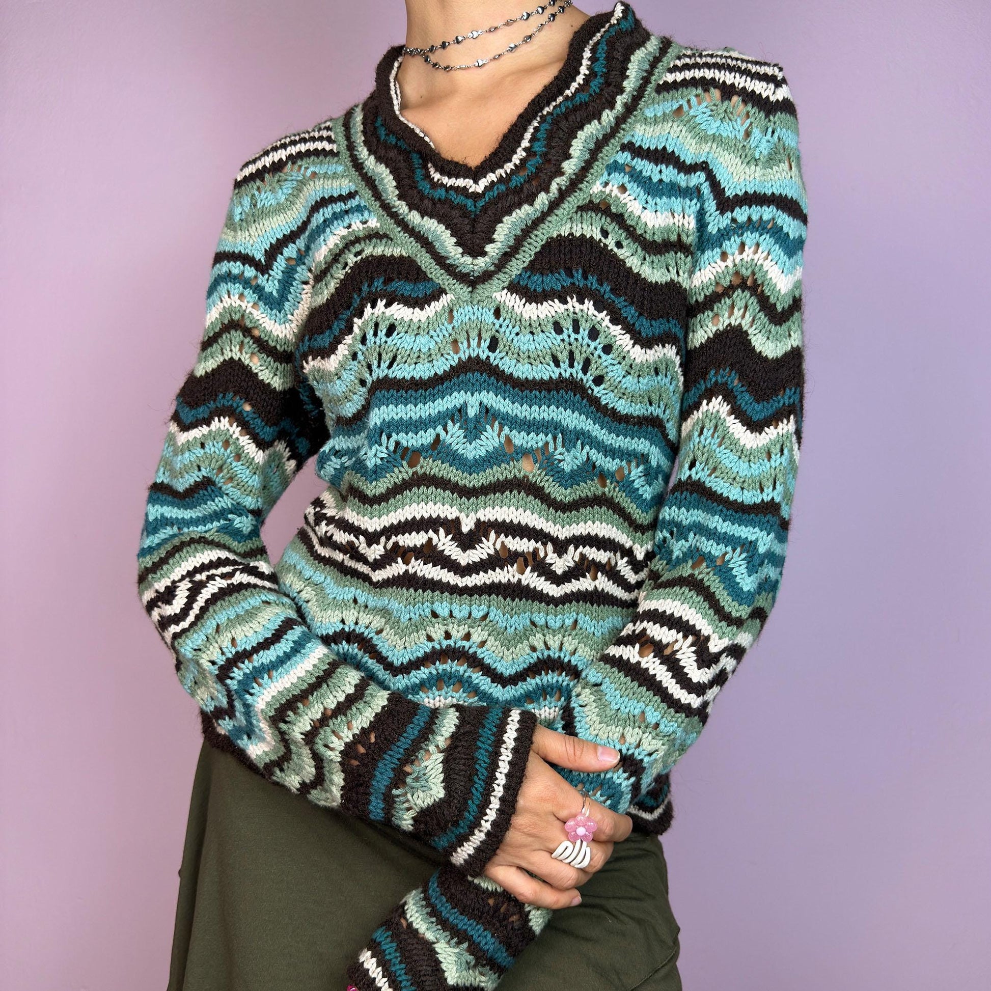 The Y2K Wave Open Knit Sweater is a vintage 2000s pullover featuring a mesmerizing wave-like pattern in shades of teal, brown and beige. This cozy sweater showcases a textured open-knit design, long sleeves, and a v-neckline. adult world shop