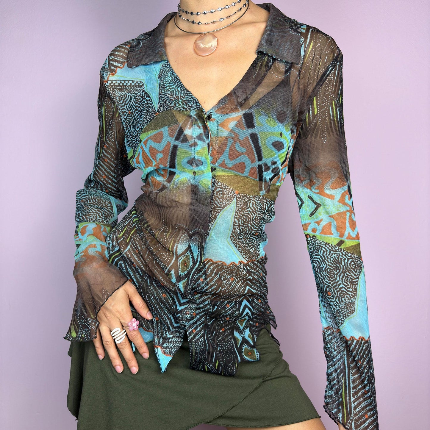 The Y2K Abstract Mesh Blouse is a vintage 2000s edgy and whimsical piece featuring a semi-sheer mesh fabric with bold, abstract patterns in earthy and blue tones and a button-front closure. The long flared sleeves. adult world shop