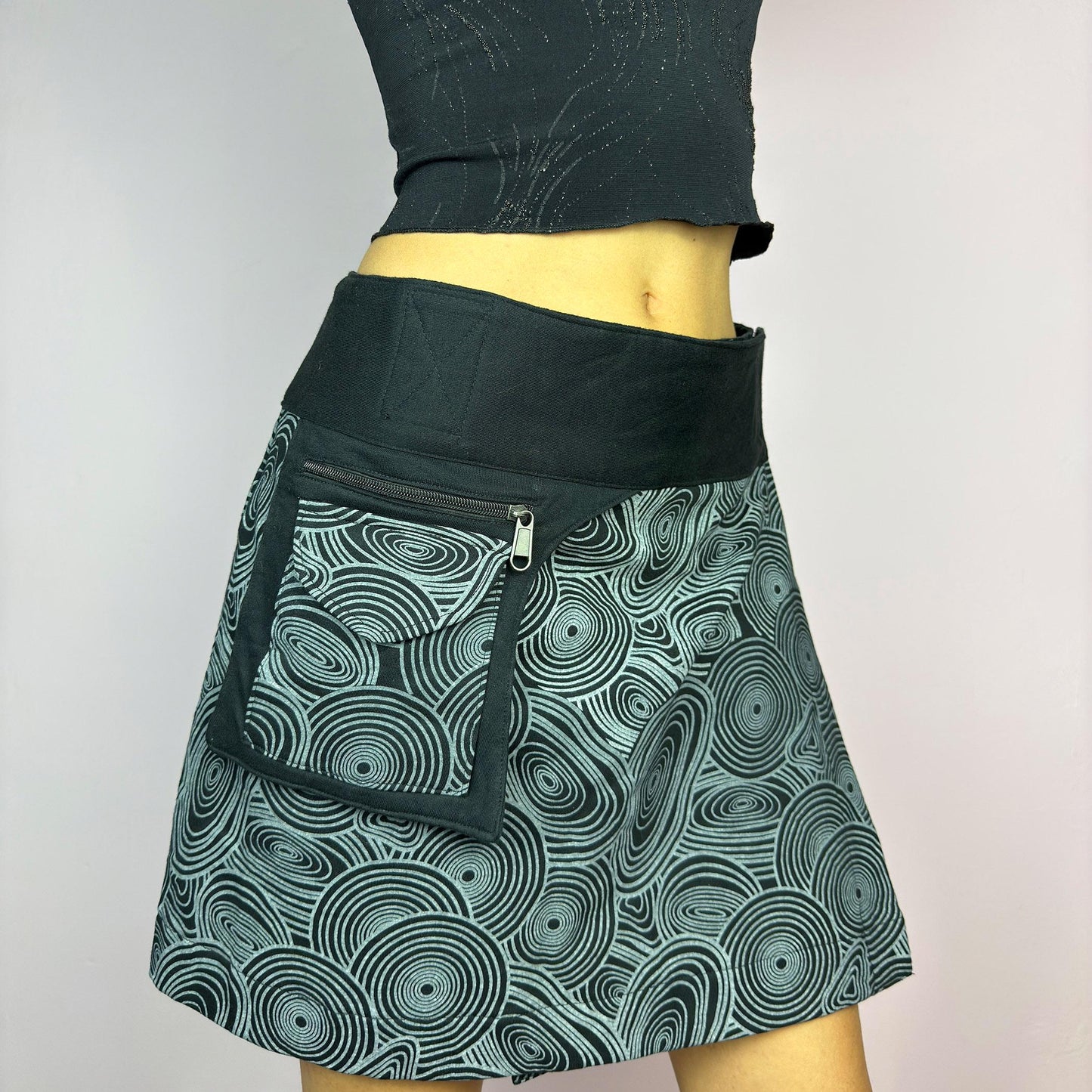 The Y2K Wrap Black Mini Skirt is a vintage 2000s piece designed with a Velcro wrap closure for an adjustable fit. Made from a thick fabric, it features a bold geometric swirl print and a utility pocket with a zipper closure. adult world shop