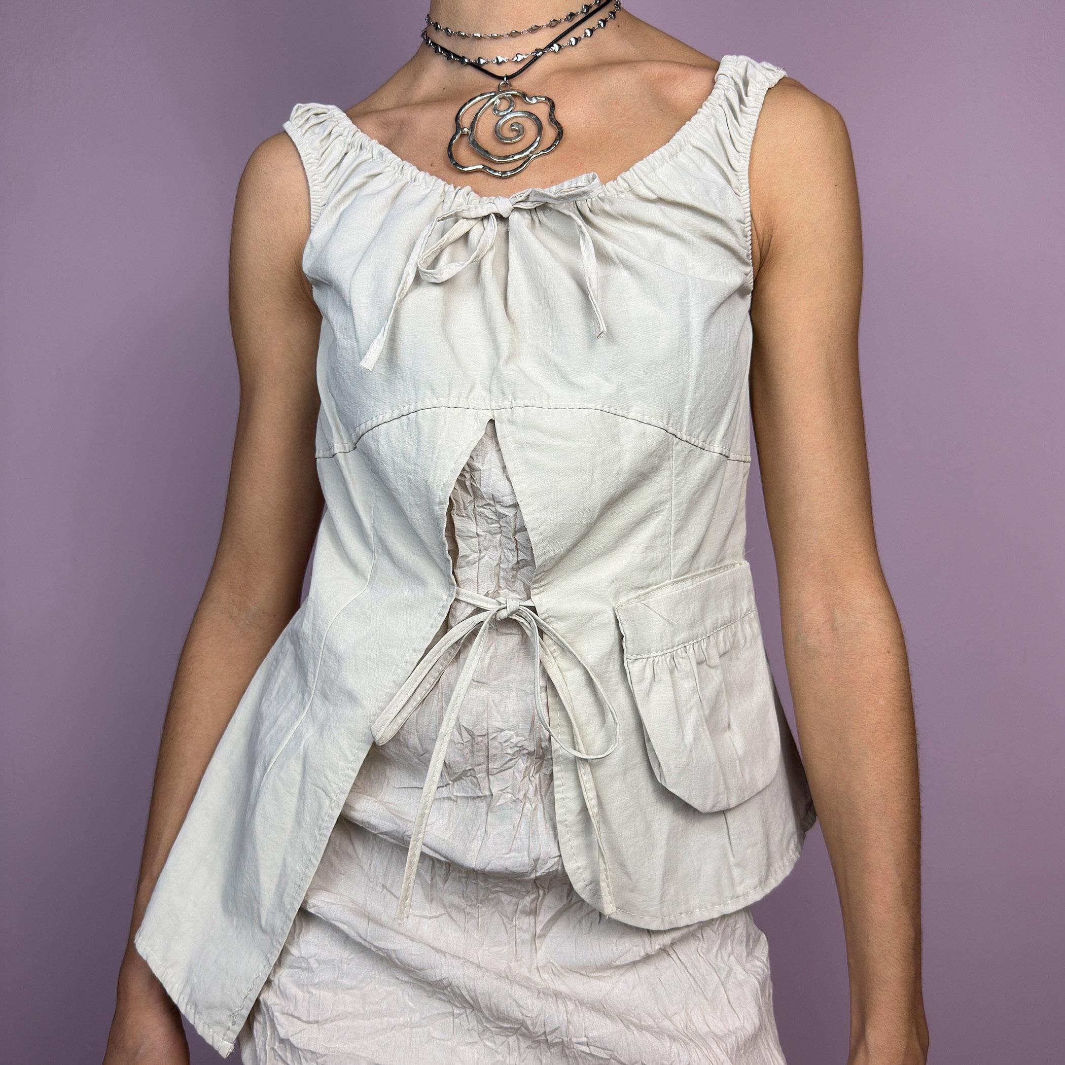 The Y2K Cream Asymmetric Tie Top is a vintage 2000s piece with a deconstructed aesthetic, featuring an asymmetric cut with a ruched neckline and delicate tie detail. The split front with adjustable ties creates a layered effect. adult world shop